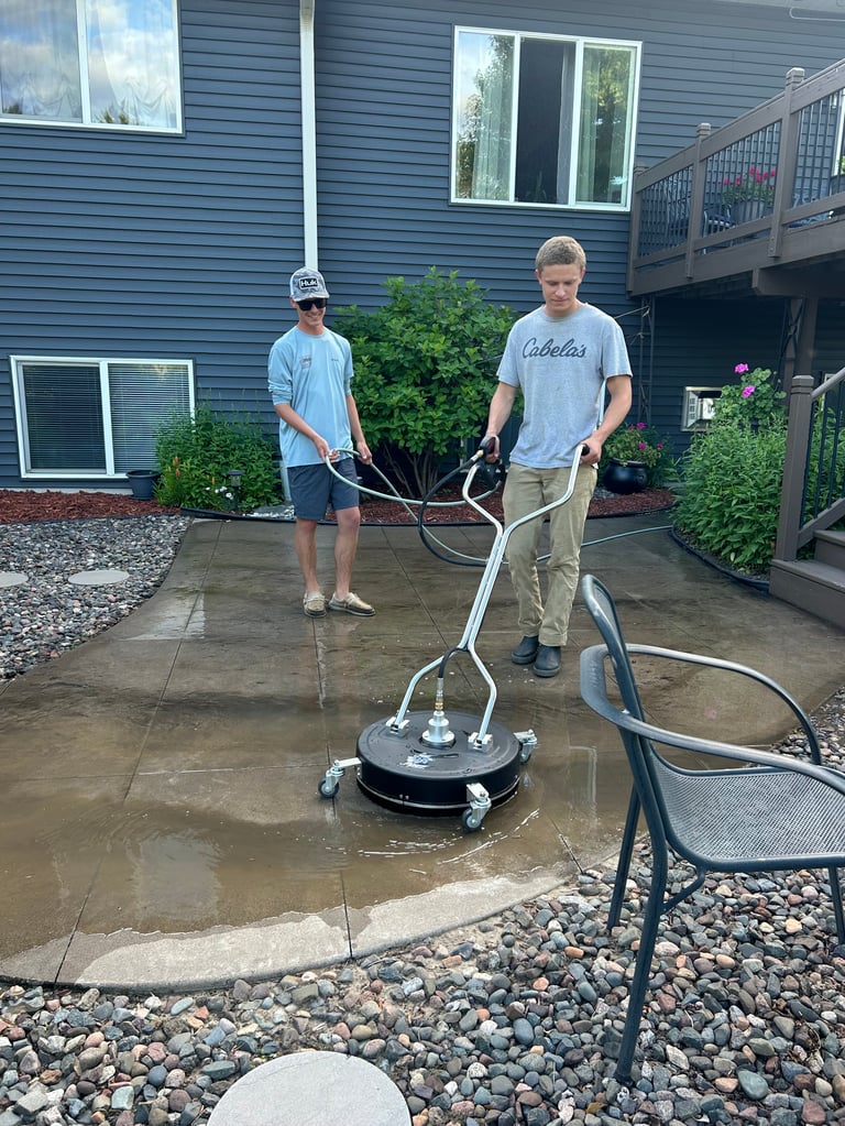 Two men pressure washing a wet patio with a power washing machine in front of a blue house