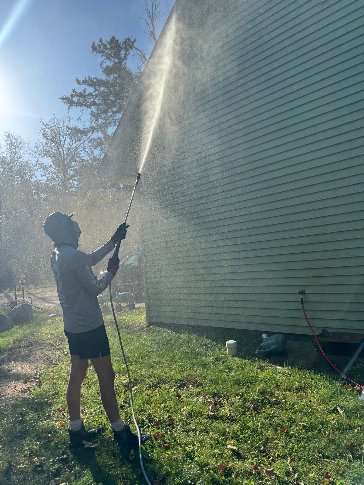 Person using pressure washer to clean side of green vinyl-sided house on sunny day